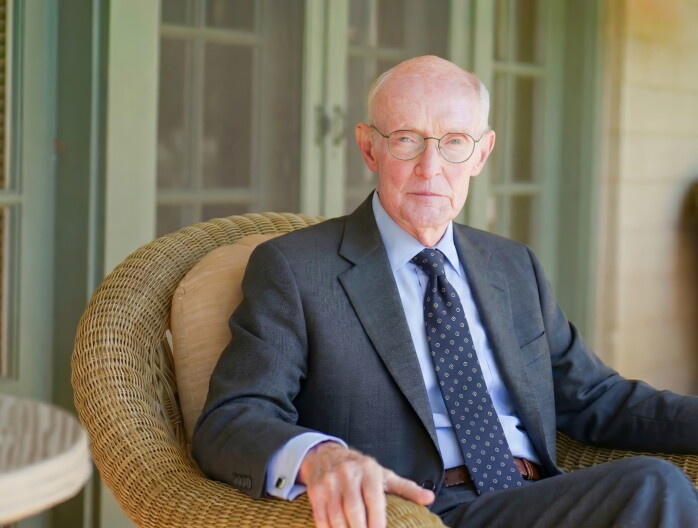 A portrait of an older white man in a gray suit. He's wearing glasses and sitting in a wicker-style chair while facing the camera.