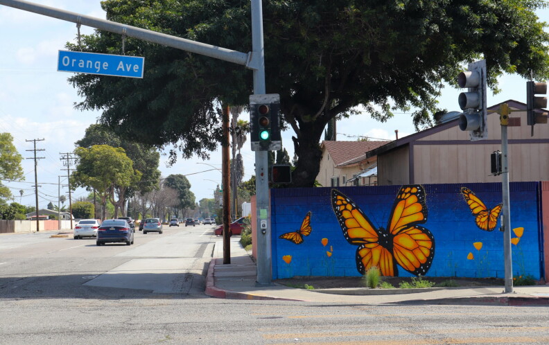A mural painted on the side of a hollow block fence depicts three orange and black monarch butterflies flying above orange poppies. The mural its on a street corner with a street sign that reads, "Orange Ave."